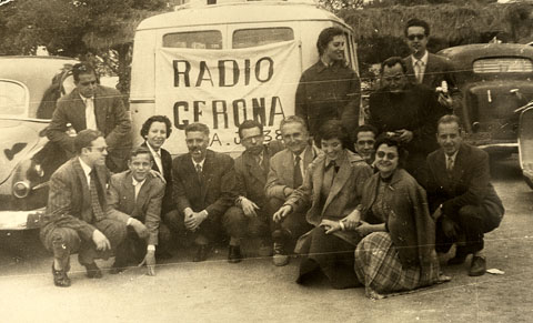 Celebració de la festa de l'Arcàngel Gabriel, patró dels radiofonistes. Retrat de grup dels treballadors de Radio Gerona EAJ38 davant d'una furgoneta amb una pancarta. S'identifiquen, ajupits, el primer a l'esquerra, Jaume Teixidor, la tercera, Francina Boris i el quart, Emilio Banda. 1955
