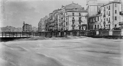 Vista del riu Onyar durant l'aiguat de l'any quaranta on apareix el pont del Pes de la Palla superat per l'aigua. A la dreta s'observa la rambla Verdaguer i algunes persones amb paraigües contemplant el riu. 1940