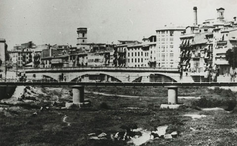 El riu Onyar amb el pont del Pes de la Palla i el pont de Pedra. En primer terme es veuen unes dones rentant al riu. Al fons, el campanar de l'església del Mercadal, la torre de les Aigües i la xemeneia de la Gròber. 1926-1935