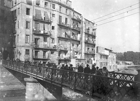 Gent creuant el pont de Sant Agustí, sobre el riu Onyar. Al fons, a la dreta, el pont d'en Gómez i el pont del Ferrocarril. 1915-1920