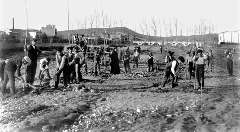 Alumnes de la granja-Escola de Sant Isidre netejant les terres de cultiu de l'escola després d'un aiguat, al barri de Pont Major. Al fons, d'esquerra a dreta, l'església parroquial de Sarrià de Ter i el pont de l'Aigua sobre el riu Ter. 1920