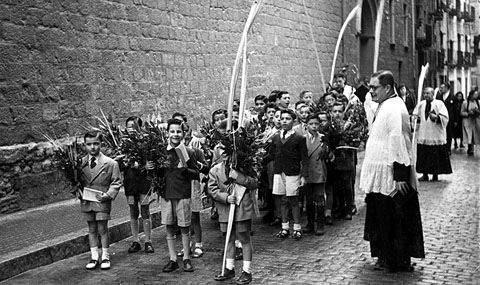 Escolania del Mercadal participant en la processó de Rams pels carrers de la ciutat. Retrat dels alumnes de l'escolania amb palmons i llorer, acompanyats per mossèn Ferran Forns, al seu pas pel carrer Obra. 1954