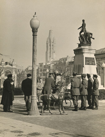 Vista la plaça de la Independència. En primer terme davant del monument s'observa un fotògraf ambulant amb un cavall de joguina envoltat per diverses persones, entre ells quatres soldats. 1957
