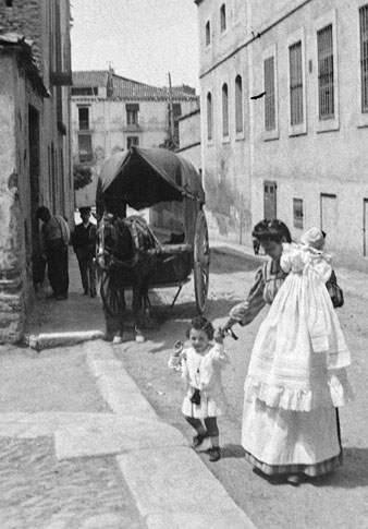 Dida passejant amb un nadó als braços i una criatura agafada de la mà en un carrer d'un lloc no identificat. Al fons, una tartana amb un cavall. 1910