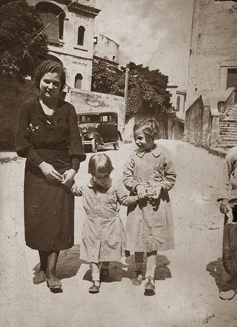 Retrat de d'una dona i dues nenes amb bata escolar al carrer del Portal Nou. A l'esquerra, la casa Gifré i a la dreta, l'església de la Mercè. 1934