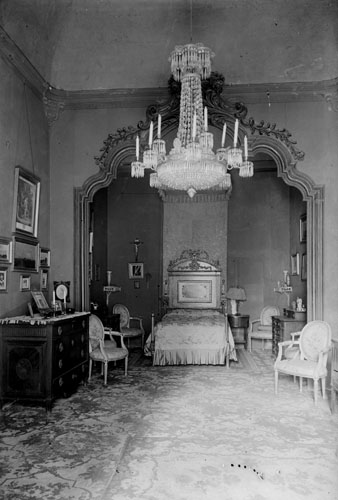 Dormitori senyorial de la casa Carles. Del mobiliari i la decoració en destaquen el llit amb capçalera, la calaixera, les cadires, els quadres i la làmpada del sostre. 1911-1915